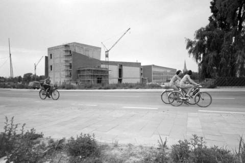 ARH NL Mellin 02-079/0010, Fahrradfahrer auf einer Straße vor einem Gebäudeneubau, im Hintergrund eine Kirche, ohne Datum