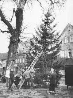 ARH Slg. Bartling 969, Männer beim Aufrichten eines Weihnachtsbaumes auf dem Kirchplatz neben der Kastanie, Blick nach Nordwesten auf die Fronten der Häuser Behrens und Knoke, Neustadt a. Rbge., um 1970