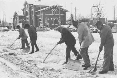 ARH Slg. Bartling 992, Fünf Männer nebeneinander beim Schneeräumen auf dem Bahnhofsvorplatz, im Hintergrund das Bahnhofsgebäude, Neustadt a. Rbge., um 1975