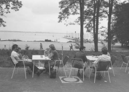 ARH Slg. Bartling 1223, Nordufer, Blick auf eine zwei Gruppen von Gästen auf der Terrasse des Restaurants Weisse Düne, dahinter der Uferweg, der leere Badestrand und der See mit Bootssteg (rechts), Steinhuder Meer, 1971
