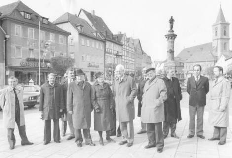 ARH Slg. Bartling 1326, Eine Gruppe von Herren auf dem Marktplatz stehend, im Hintergrund die Bürgerhäuser (links), der Marktbrunnen und die Pfarrkirche Mariä Himmelfahrt, Bad Neustadt a. d. Saale, um 1980
