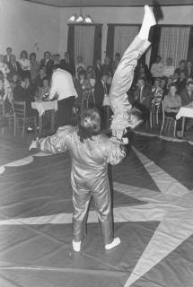 ARH Slg. Bartling 1778, Akrobatische Einlage (Handstand eines Kindes auf dem Arm eines Erwachsenen) bei einem Festball in der Bürgerhalle (?), Blick über die Akrobaten auf die Gäste am Tisch, Neustadt a. Rbge., um 1970