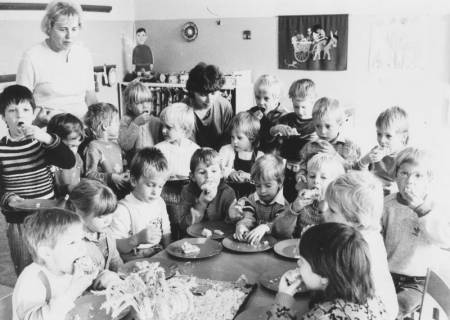ARH Slg. Bartling 1874, Gruppe von Kindern, die um einen Tisch sitzen oder stehen und einen Kuchen verzehren, mit zwei Erzieherinnen, Neustadt a. Rbge., um 1970