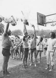 ARH Slg. Bartling 2040, Kinder auf einem Sportplatz versuchen mit einer spitzen Mütze auf dem Kopf nach Ballons, die mit Wasser gefüllt sind und auf einer Leine an einem Basketballkorb aufgereiht sind, zu springen und sie dadurch zum Platzen zu bringen, um 1975