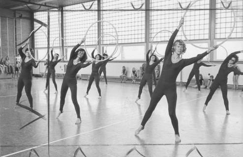 ARH Slg. Bartling 2115, Gymnastikübung einer Gruppe von älteren mit Sportleggings bekleideten Frauen, die in der TSV-Turnhalle einen Gymnastikreifen über dem Kopf wiegen, 1974