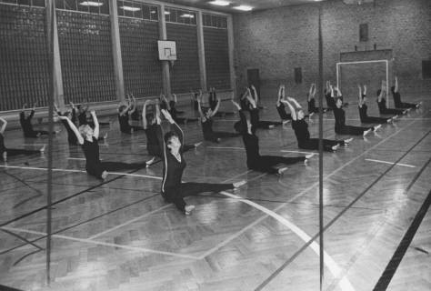 ARH Slg. Bartling 2124, Gymnastik-Übung von Frauen, die in Sportleggings auf dem Boden einer Halle (mit Glasbausteinwand) sitzen, die Arme nach oben streckend, Neustadt a. Rbge., 1972