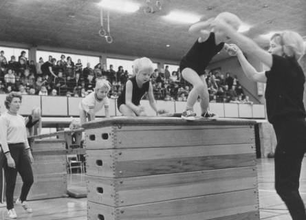 ARH Slg. Bartling 2126, Vorschul-Kinder bei einer Grundübung in einer Turnhalle unter Aufsicht von zwei erwachsenen Helferinnen hintereinander kriechend und von einem Kasten springend, Neustadt a. Rbge., 1972