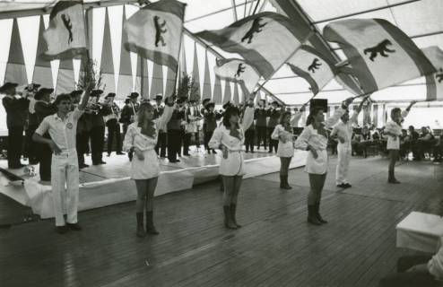 ARH Slg. Bartling 2210, Auftritt einer Gruppe von Fahnenschwenkerinnen und -schwenkern mit Berliner Flaggen in einem Festzelt, im Hintergrund ein Trompeter- und Showcorps, um 1970