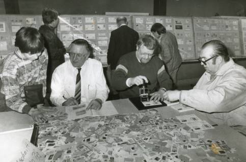 ARH Slg. Bartling 2259, Drei Philatelisten (l. Heinrich Schöneberg-Schnappka) an einem Tisch sitzend, daneben ein junger Briefmarkenfreund, die auf dem Tisch ausgebreitete Schnittmarken sortieren, im Hintergrund die aufgestellten Ausstellungswände mit Exponaten, Neustadt a. Rbg., 1973