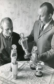 ARH Slg. Bartling 2292, Der 1. Vorsitzende der Sportschützen Fritz Jonat (r.) und N. N. begutachten fünf Pokale auf einem runden Tisch mit Häkeldecke, Neustadt a. Rbge., 1974