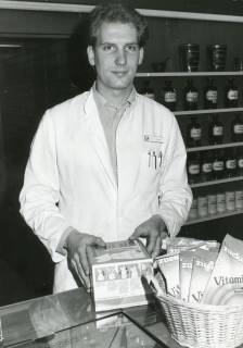 ARH Slg. Bartling 2412, Apotheker Dietrich Redeker hinter dem Tresen stehend, im Hintergrund ein Regal mit gläsernen Behältnissen, Neustadt a. Rbge., um 1970