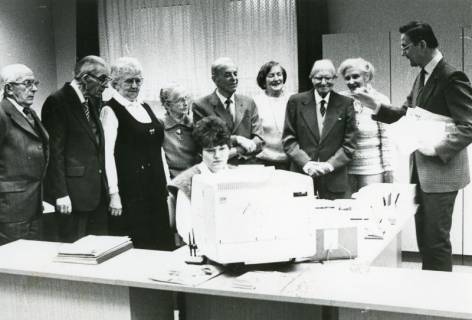 ARH Slg. Bartling 2419, Eine junge Frau am PC sitzend und schreibend, dahinter im Halbkreis stehend eine Gruppe von Männern und Frauen, die dem Leiter der Barmer Ersatzkasse N. N. (r.) zuhören, Neustadt a. Rbge., um 1980