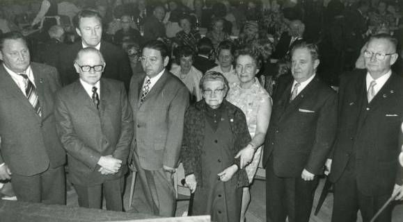 ARH Slg. Bartling 2429, Sechs Männer und zwei Frauen vom Reichsbund nebeneinander stehend vor dem an Tischen sitzenden Publikum, Garbsen, 1973