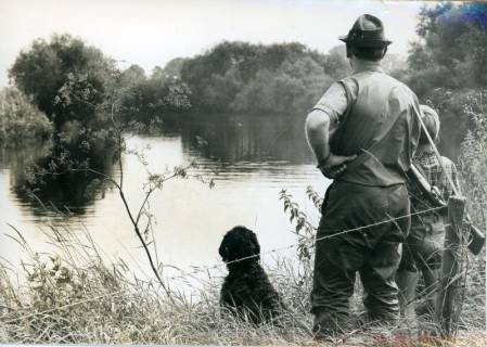 ARH Slg. Bartling 2473, Jagdpächter Dr. med. dent. Henning Gode mit Hut und geschulterter Flinte, begleitet von einem Jungen und einem Jagdhund am Ufer eines Sees stehend und auf das Wasser schauend, Neustadt a. Rbge., um 1970