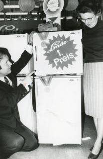 ARH Slg. Bartling 2517, Elektro-Händler Ulrich Pallak kniet links neben einem Kühlschrank der Firma Linde und zeigt auf ein am Kühlschrank befestigtes Werbeschild mit der Aufschrift "Linde, 1. Preis", Neustadt a. Rbge., 1972