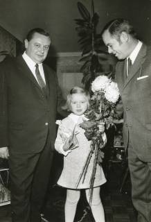 ARH Slg. Bartling 2580, Mädchen mit einem Strauß Chrysanthemen in der Linken und einer prall gefüllten Tüte in der Rechten zwischen zwei mit Anzügen bekleideten Herre in einem Wohnzimmer mit Gummibaum im Hintergrund, Neustadt a. Rbge., um 1975