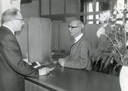 ARH Slg. Bartling 2756, Einzahlung eines Geldbetrages durch einen Kunden an Angestellten hinter dem mit Blumen geschmückten Tresen eines Schalters, Neustadt a. Rbge., 1969