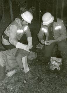ARH Slg. Bartling 3413, Erste-Hilfe-Übung: Zwei Feuerwehrleute versorgen einen bewusstlosen auf dem Waldboden liegenden Mann mit stark blutender Kopfwunde (laut Beschriftung eines um den Hals gehängten Schildes), Neustadt a. Rbge., um 1974