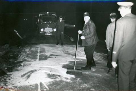 ARH Slg. Bartling 3435, Unter den Augen eines Polizisten (r.) zwei Feuerwehrleute im Dunkeln beim Fegen eines aufgestreuten Bindemittels auf der Straße (im Hintergrund ein Kleinlaster der Feuerwehr), Neustadt a. Rbge., 1970