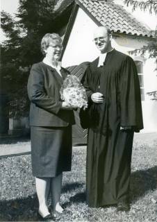 ARH Slg. Bartling 3462, Pastor N. N. mit Talar und Beffchen auf dem Rasen stehend neben seiner Frau (?) mit Blumenstrauß, Neustadt a. Rbge., um 1975