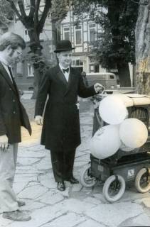 ARH Slg. Bartling 3493, Auftritt eines Leierkastenmanns mit Frack und Bowler auf dem Kirchplatz vor der Liebfrauenkirche, am Leierkasten drei Luftballons, links daneben stehend ein Mann, Neustadt a. Rbge., um 1975