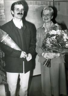 ARH Slg. Bartling 3667, Lehrerin N. N. (mit Blumenstrauß im linken Arm) neben Lehrer N. N. (mit Einschulungstüte im rechten Arm) vor einer Stundentafel stehend, Neustadt a. Rbge., 1987