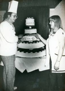 ARH Slg. Bartling 3735, Vierstöckige Torte mit der Aufschrift ".WK 10 Jahre", links daneben ein Koch in Berufskleidung, rechts eine Angestellte in weißem Kittel, Schneeren, um 1980