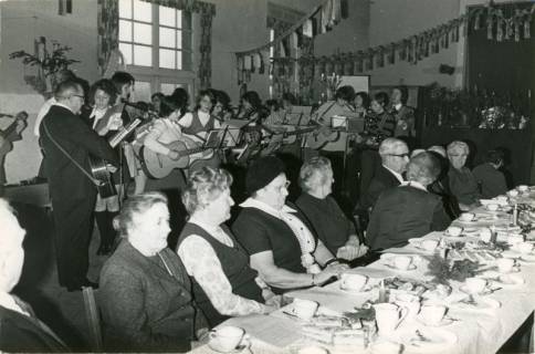 ARH Slg. Bartling 3855, Adventliche Kaffeetafel für Senioren, Blick über die Leute am gedeckten Tisch auf eine Gruppe junger Leute mit Gitarren, Bordenau, 1972