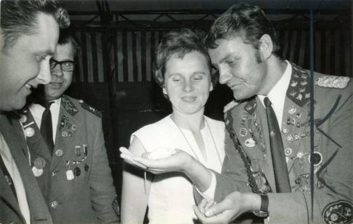 ARH Slg. Bartling 3867, Ein uniformierter Schütze zeigt (unter den Augen zweier Kameraden) einer jungen Frau eine weiße Maus in seiner flachen Hand beim Schützenfest, Bordenau, 1971