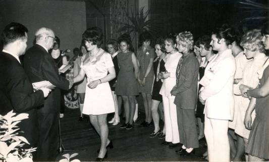 ARH Slg. Bartling 4164, Überreichung eines Dokuments in einem Saal an eine junge Frau durch den N. N., hinter ihr zahlreiche andere Frauen in Wartestellung, Empede (?), 1970