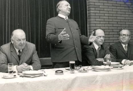 ARH Slg. Bartling 4195, Henry Hahn in Trachtenjacke, vor einer Theaterbühne stehend und mit ausgebreiteten Händen redend, an einem zum Essen gedeckten Vorstandstisch zwischen dem sitzenden Bürgermeister Mehring, Berenbostel (l.) und N. N. sowie N. N., Hagen, 1973