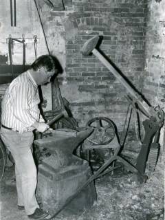 ARH Slg. Bartling 4263, Vorsitzender des Schmiedevereins Helstorf N. N. in Freizeitkleidung beim Versuch, mit dem Hammer am Amboss ein Stück Eisen zu formen in der Alten Schmiede, Helstorf, um 1980