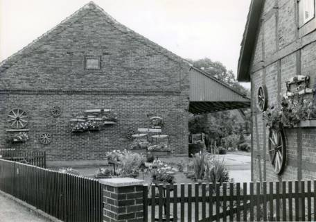 ARH Slg. Bartling 4303, Vorgarten an einem Bauernhof dessen Außenwände mit alten Wagenrädern und hölzernen Wagendeichseln behängt sind, Luttmersen, um 1980