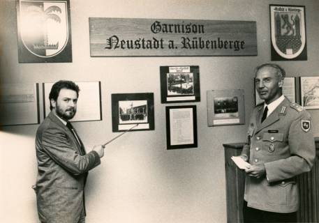 ARH Slg. Bartling 4333, Oberstleutnant Streibel (r.) und ein Zivilist vor einer Wand mit historischen Fotos und Wappen sowie einem Wandschild mit dem Schriftzug "Garnison Neustadt a. Rübernberge" stehend, Luttmersen, um 1985