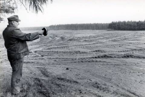 ARH Slg. Bartling 4414, Der Meteler Ortsbürgermeister Heinrich Dettmering mit einem Pilz in der ausgestreckten Hand stehend am Rande des Standortübungsgeländes, Blick über den Platz, der von Spuren der Kettenfahrzeuge gekennzeichnet ist, Luttmersen, um 1980