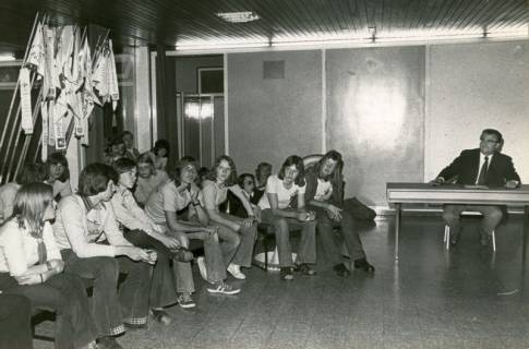 ARH Slg. Bartling 4575, Jugendliche im DRK-Heim unter Wimpeln in einer Reihe sitzend in Diskussion mit einem Mann, der am Tisch sitzt, Mardorf, 1973