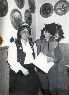 ARH Slg. Bartling 4610, Aufführung des Theaterclubs im Schützenhaus, Blick auf eine Szene mit zwei Frauen (eine mit Diadem einer Schützenkönigin, die andere verkleidet als Mann), Kniestück, an der Wand Schützenscheiben von Schützenköniginnen, Mardorf, um 1980