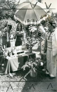 ARH Slg. Bartling 4651, Präsentation von Modeschmuck durch zwei Teenager unter einem Sonnenschirm an einer Parkbank auf einem Basar, rechts die Lehrerin Frau Kreye, Metel, um 1975