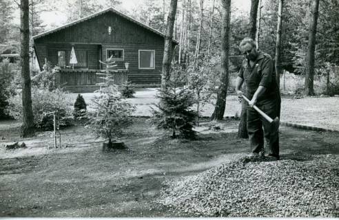 ARH Slg. Bartling 4657, Wochenendhaus (Blockhaus) in Kiefernwaldlichtung, Außenansicht Giebelseite, rechts ein Mann bei Verrichtung gärtnerischer Arbeiten, Metel, 1975
