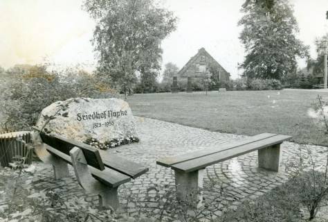 ARH Slg. Bartling 4738, Gedenkstein (Findling) am Friedhof Nöpke 1873-1955, Blick über das eingeebnete Areal auf drei übrig gebliebene Steinsäulen und den Giebel eines am Rande stehenden Hauses (2 Ex.), Nöpke, um 1975