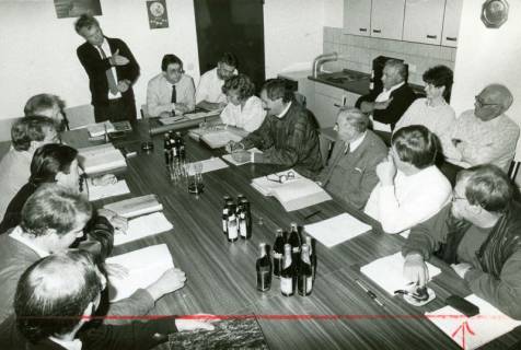 ARH Slg. Bartling 4932, Ortsbürgermeister von Scharrel Wilfried Meißner (l.) am Kopfende eines Tisches stehend und die Ortsratsmitglieder am Tisch sitzend in einem Raum, der mit einer Küchenzeile ausgestattet ist, Scharrel, um 1975