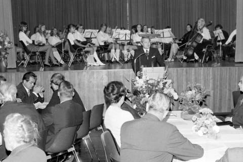 ARH Slg. Weber 02-024/0009, Ein Mann hält an einem Podium eine Rede, im Hintergrund ein Orchester auf einer Bühne, in der Festhalle, Gehrden, zwischen 1965/1970