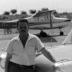 Archiv der Region Hannover, ARH NL Mellin 01-088/0002, An ein Auto lehnender Mann mit einem Motorflugzeug im Hintergrund