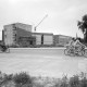 Archiv der Region Hannover, ARH NL Mellin 02-079/0010, Fahrradfahrer auf einer Straße vor einem Gebäudeneubau, im Hintergrund eine Kirche