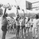 Stadtarchiv Neustadt a. Rbge., ARH Slg. Bartling 2040, Kinder auf einem Sportplatz versuchen mit einer spitzen Mütze auf dem Kopf nach Ballons, die mit Wasser gefüllt sind und auf einer Leine an einem Basketballkorb aufgereiht sind, zu springen und sie dadurch zum Platzen zu bringen
