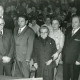 ARH Slg. Bartling 2429, Sechs Männer und zwei Frauen vom Reichsbund nebeneinander stehend vor dem an Tischen sitzenden Publikum, Garbsen