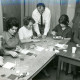 ARH Slg. Bartling 3045, Kreisberufsschule, Schulungsraum, fünf jüngere Frauen an Übereck-Tischen sitzend bei Mosaikarbeiten mit Perlen (?) unter der Aufsicht eines Mannes, Neustadt a. Rbge.