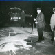 Stadtarchiv Neustadt a. Rbge., ARH Slg. Bartling 3435, Unter den Augen eines Polizisten (r.) zwei Feuerwehrleute im Dunkeln beim Fegen eines aufgestreuten Bindemittels auf der Straße (im Hintergrund ein Kleinlaster der Feuerwehr), Neustadt a. Rbge.