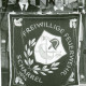 Stadtarchiv Neustadt a. Rbge., ARH Slg. Bartling 3730, Fahne der Freiwilligen Feuerwehr Scharrel 1954, ausgebreitet gehalten von zwei Feuerwehrleuten, im Hintergrund ein Regal mit der Montur, Scharrel