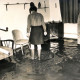 Stadtarchiv Neustadt a. Rbge., ARH Slg. Bartling 4243, Überschwemmung im Neubaugebiet, Frau mit Stiefeln geht durch das Wasser in ihrem Wohnzimmer, Helstorf